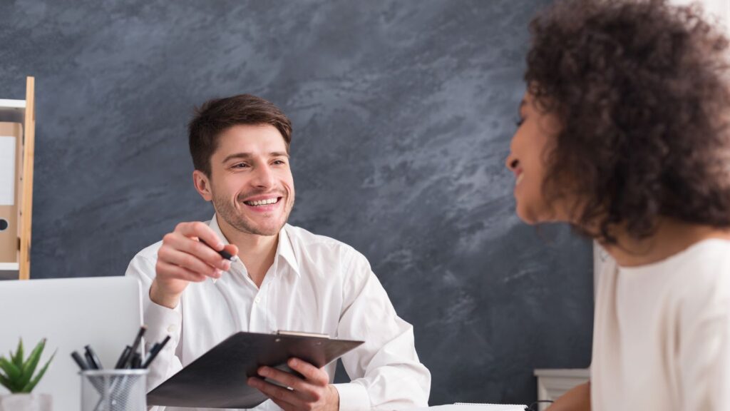Deeskalierende Feedbackgespräche: Eine Führungskraft mit einem Klemmbrett im Gespräch mit einer Mitarbeiterin, beide lächeln zufrieden